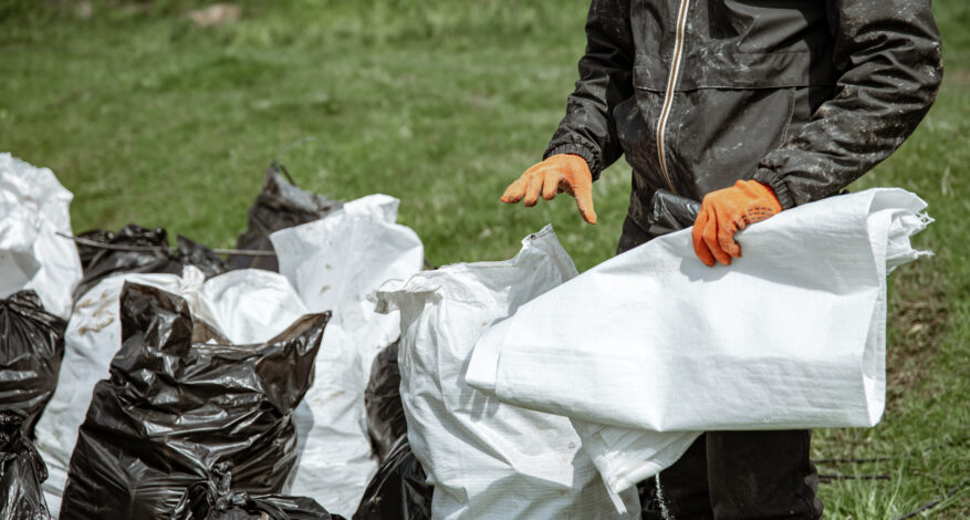 Worker in orange gloves holding a woven polypropylene geotextile bag beside filled sand bags